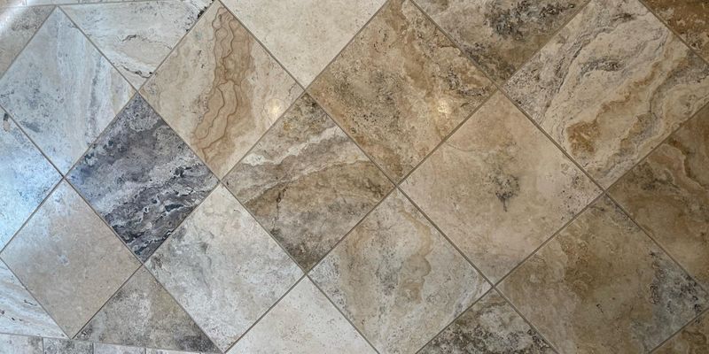 An overhead view of a floor with natural stone tiles in shades of beige, grey, and cream, laid in a diamond pattern with clean, uniform grout lines.
