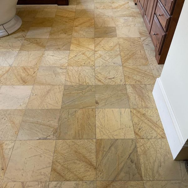 A bathroom floor with square, golden-yellow stone tiles that have a textured, veined appearance, shown next to a dark wood vanity and a white wall.