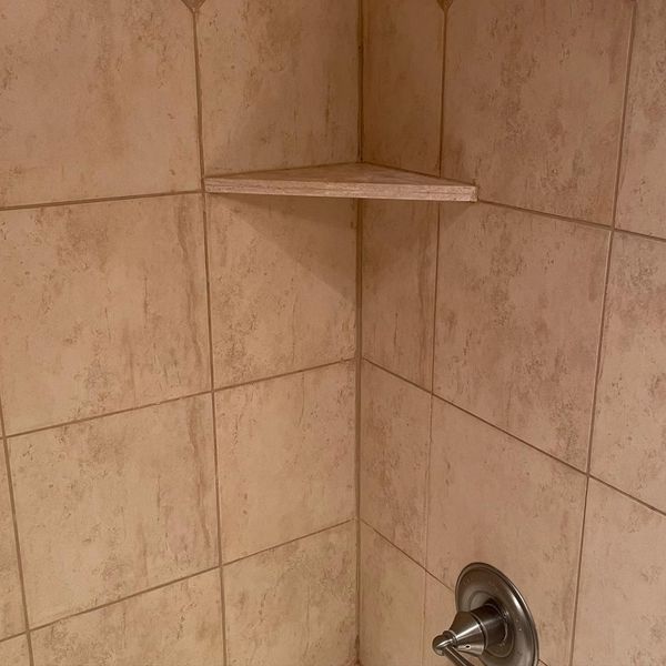 The inside corner of a shower with large, square, beige-colored tiles, a matching triangular corner shelf, and a silver shower handle, all with clean grout.