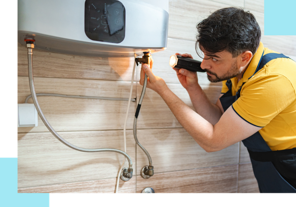 A plumber in a yellow shirt and dark overalls using a flashlight to inspect the bottom pipe connections of a tankless water heater.