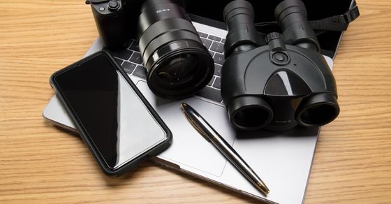 a phone, pen, binoculars, camera, and a laptop on a desk