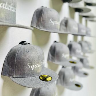 Baseball caps displayed front-facing on a wall featuring The Original Squatchee™ brand using patented adhesive hat hooks. Statement Baseball Cap Display