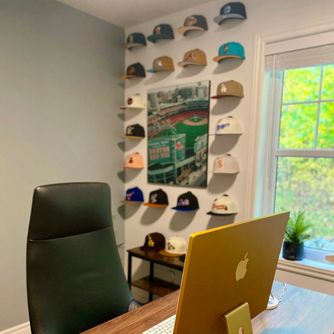 Baseball caps displayed front-facing in a home office to create a professional backdrop for virtual meetings using The Original Squatchee™ adhesive hat hooks. Home Office & Virtual Meeting Hat Wall