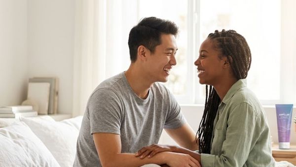 A happy young couple sharing an intimate moment in a bright, modern bedroom with a premium lubricant bottle on the nightstand.