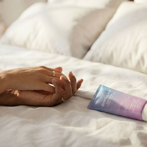 Two people holding hands on white bedsheets next to a lubricant bottle representing comfort and intimacy.