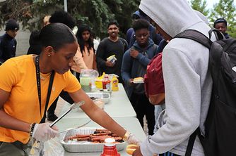 Youth outreach worker serves hotdogs to teenage boy. A line of teenagers stand behind ready to receive a hotdog also.