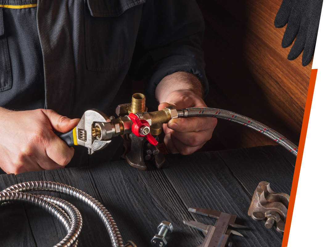 A plumber's hands using an adjustable wrench to tighten a brass valve assembly connected to a flexible braided hose on a dark wooden workspace.