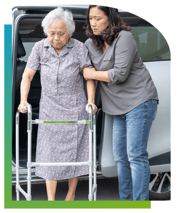A caregiver helping an elderly woman step out of a car while using a walker.