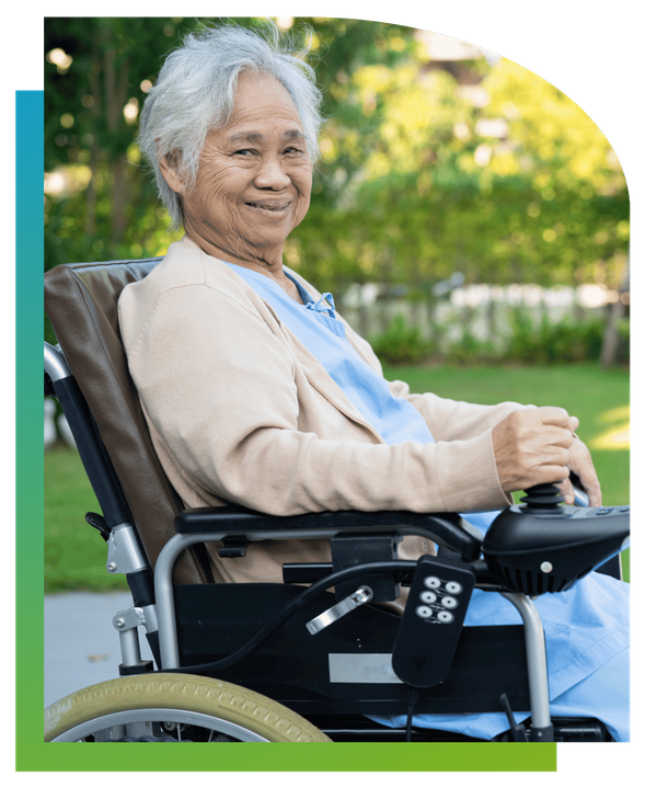 A smiling elderly woman sitting in a wheelchair outdoors on a sunny day.