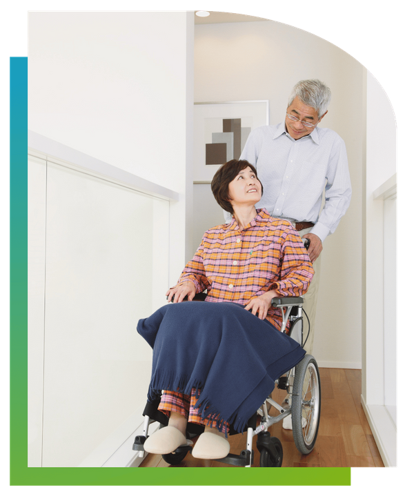 An elderly woman sitting in a wheelchair indoors, looking up and smiling at a male caregiver standing behind her.
