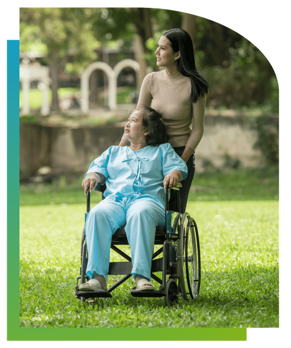 A caregiver pushing an elderly woman in a wheelchair through a sunny park.