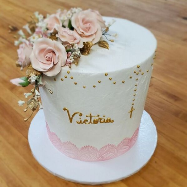 A white frosted cake with pink and gold floral decorations, gold lettering "Victoria", and a small gold cross. A white frosted cake with pink and gold floral decorations, gold lettering "Victoria", and a small gold cross.