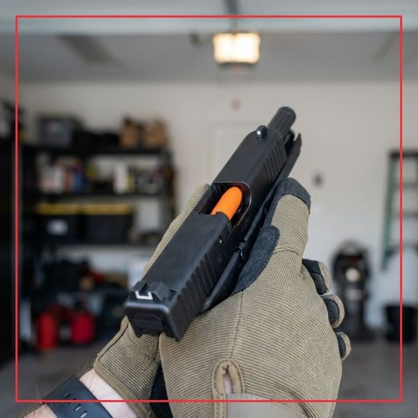 A 1:1 square photograph showing a person's hands in a garage, safely practicing dry-fire trigger control with a handgun that has a bright orange dummy round clearly visible in the open chamber