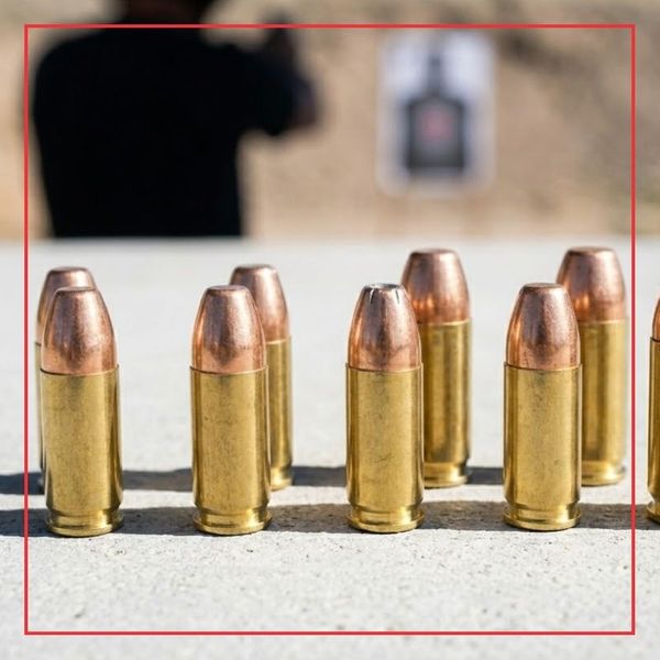 A square, sunlit photograph capturing loose brass handgun cartridges standing upright on a concrete surface at an outdoor shooting range, with a blurred silhouette of a shooter in the distance.