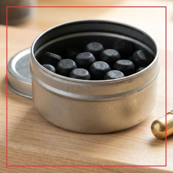 A square 1:1 photograph in a workshop setting showing an open, unmarked metal tin containing several pre-formed black powder propellant pellets on a wooden loading bench surface.