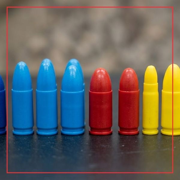 A 1:1 square macro photograph displaying a precision arrangement of colorful inert dummy rounds (blue, red, yellow) in different calibers, lined up on a dark workbench.
