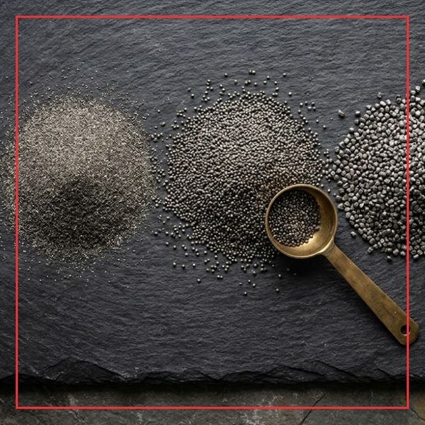 A close-up, top-down photograph in a square 1:1 format showing three distinct granulations of black powder placed on a rugged slate surface next to an aged brass measuring scoop.