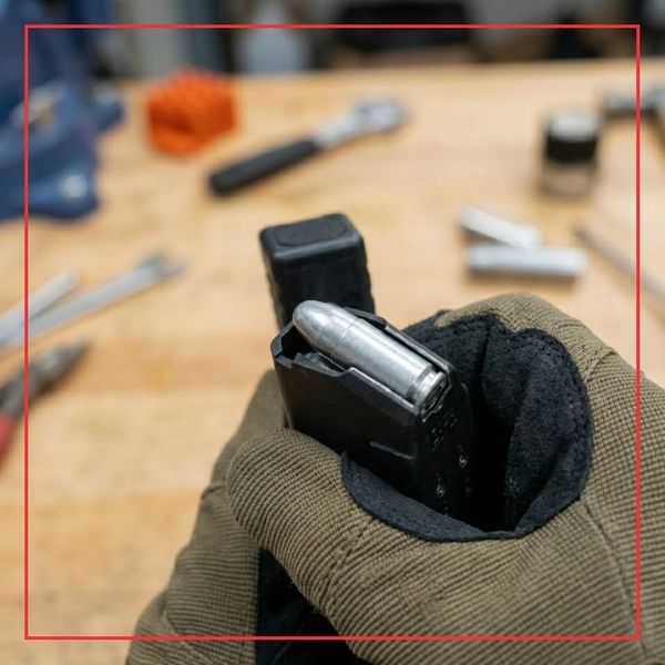 A 1:1 square photograph capturing a close-up of a gloved hand inserting a weighted, metallic silver dummy round into a pistol magazine in a workshop setting.