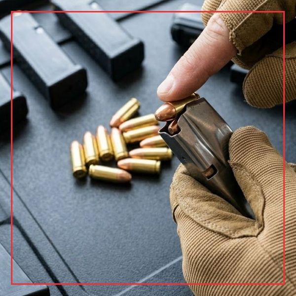 A square photograph capturing gloved hands efficiently loading a pistol magazine with multiple loose, gleaming centerfire cartridges on a workbench.