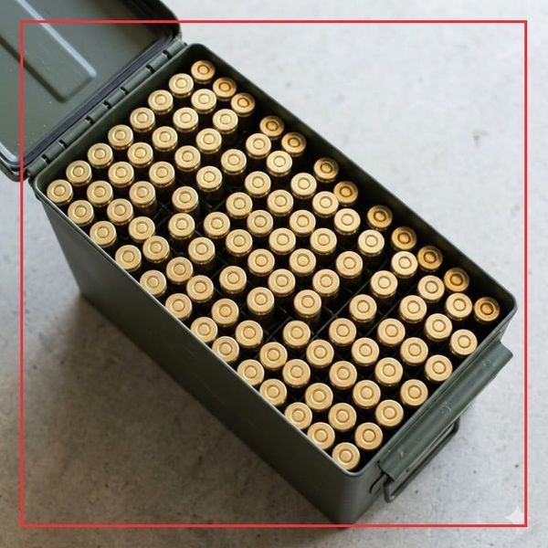 An overhead square photograph of numerous uniform, shiny brass blank cartridges organized tightly within a generic green ammunition can, signifying bulk supply.