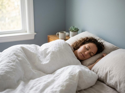 A woman with curly hair sleeps soundly on her side in a bed with white linen bedding, next to a window and a nightstand. Woman sleeping peacefully in bed with white linen