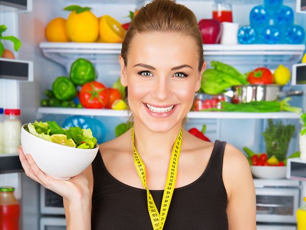 a woman holding a salad with an open refrigerator with a measuring tape around her neck 