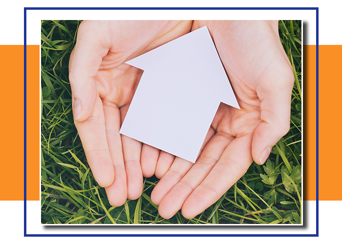 Two female hands holding a small paper family house cutout over fresh green grass.