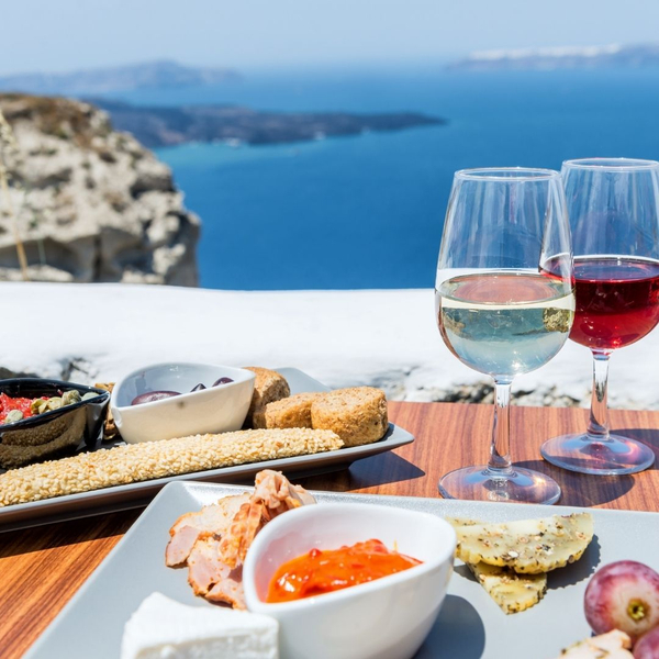 An image of two wine glasses and plates of Greek Food.