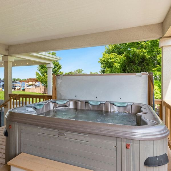 Hot tub with its cover partially opened by a cover lifter, located on a wooden deck under a covered patio with a view of a lake. Hot tub with its cover partially opened by a cover lifter, located on a wooden deck under a covered patio with a view of a lake.